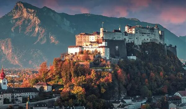 View of Hohensalzburg Fortress on a hill overlooking the city of Salzburg, surrounded by autumn-colored trees and mountains in the background.