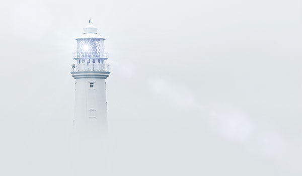 Weißer Leuchtturm mit strahlendem Licht, der aus dichtem Nebel herausragt.