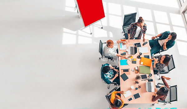 Gruppe von Menschen sitzt an einem Konferenztisch mit Laptops, Tablets und Notizblöcken, während im Hintergrund ein rotes Flipchart steht.