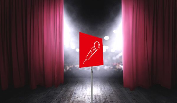 Stilisiertes Mikrofon-Symbol auf einer roten geometrischen Fläche auf einer Bühne zwischen geöffneten roten Vorhängen mit Scheinwerferlicht im Hintergrund.