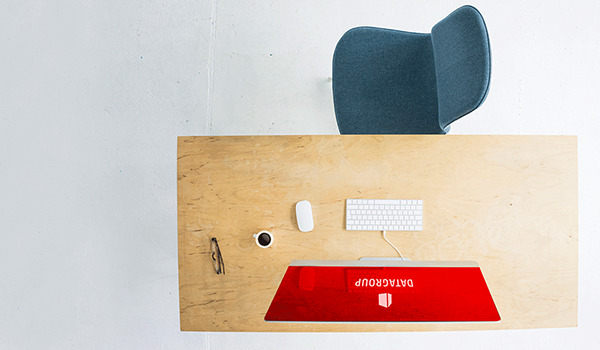 Ein minimalistischer Arbeitsplatz mit Holzschreibtisch, blauem Stuhl, weißer Tastatur. Der Bildschirm zeigt ein rotes Hintergrundbild mit dem DATAGROUP Logo.
