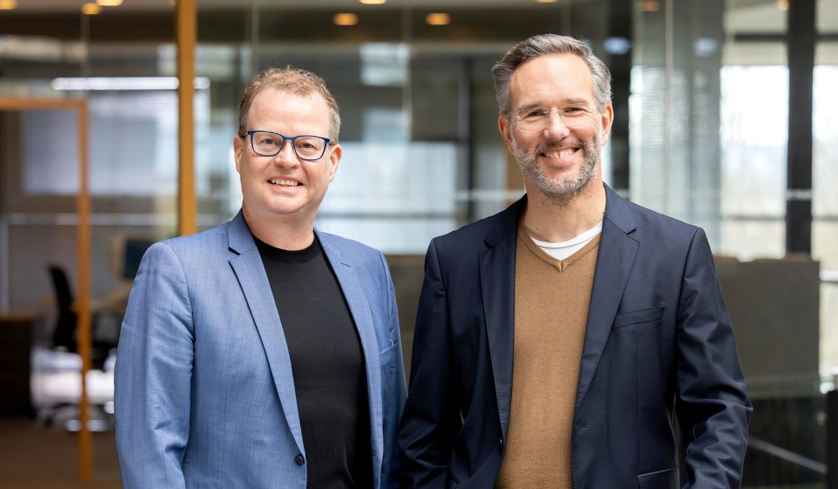 Two men in suits standing side by side in a modern office, smiling at the camera.