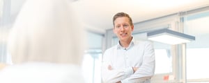 Mann im weißen Hemd steht mit verschränkten Armen in einem modernen Büro und lächelt freundlich