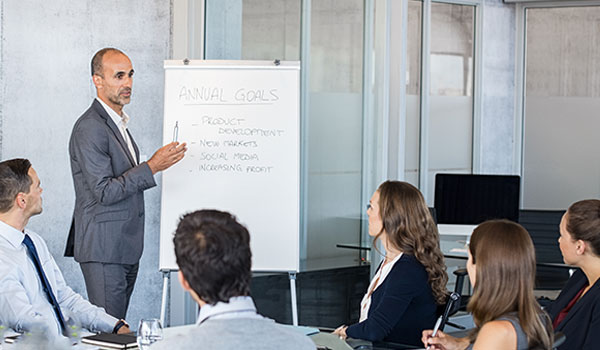 Geschäftstreffen in einem modernen Büro, bei dem ein Mann einer Gruppe von Kollegen, die um einen Tisch herum sitzen, die Jahresziele auf einem Flipchart präsentiert.