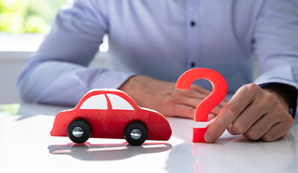 Person im Hemd, sitzt am Tisch und hält ein rotes Fragezeichen neben einem roten Spielzeugauto. Man sieht nicht das Gesicht.