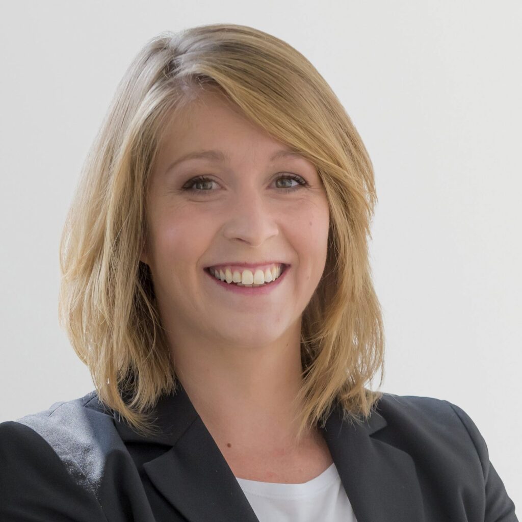 Frau mit blonden Haaren und lächelndem Gesicht, trägt einen schwarzen Blazer und steht vor einem hellen Hintergrund.